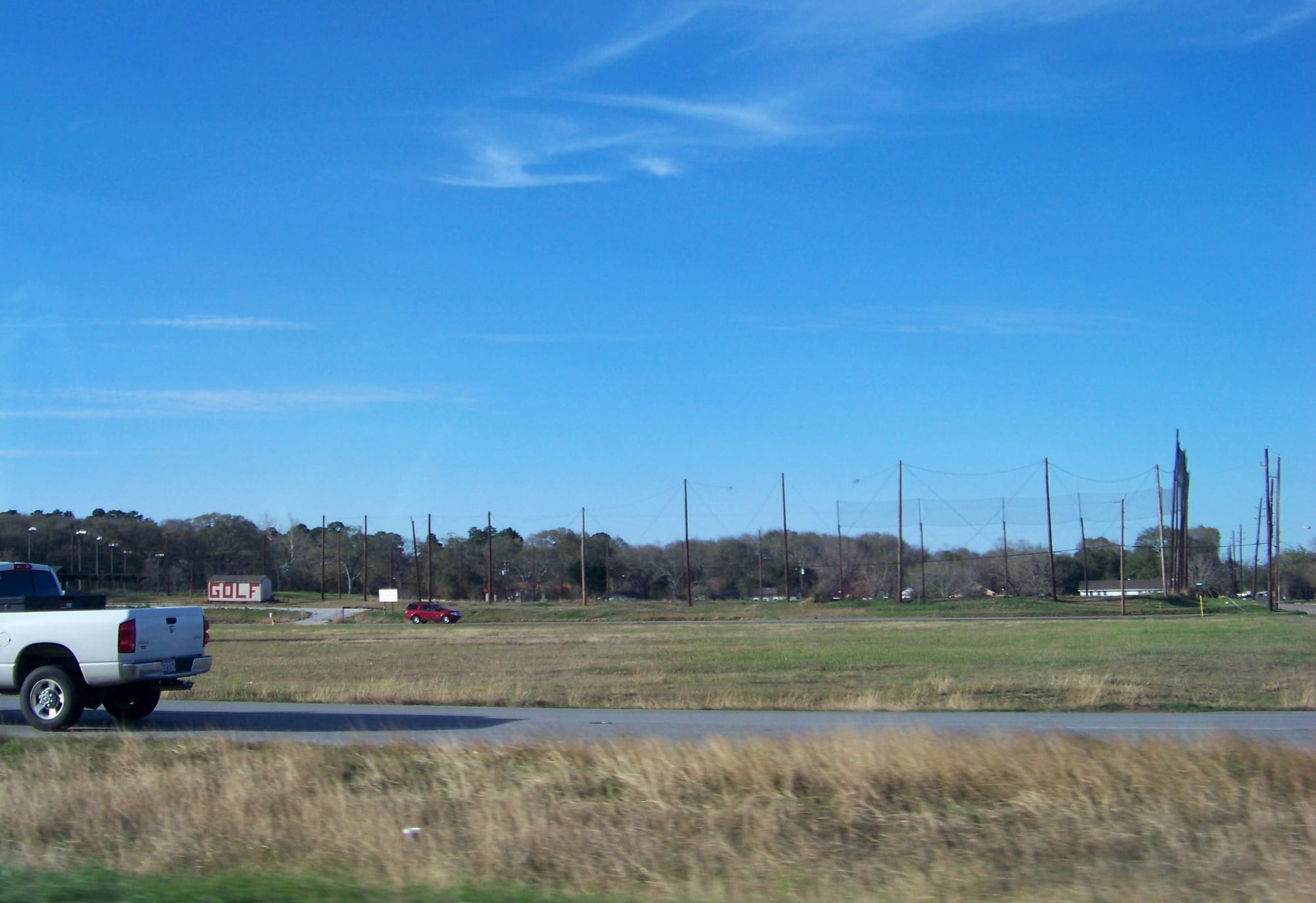 Open grassland beside a two-lane road in Katy, Texas, with a pickup truck, a tree line and a golf driving range net under a blue sky