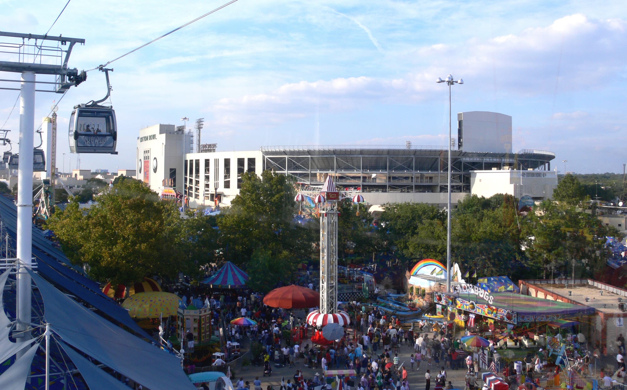 Cotton Bowl Stadium rising behind the State Fair of Texas midway, seen from the fairgrounds skyway with crowds and rides below